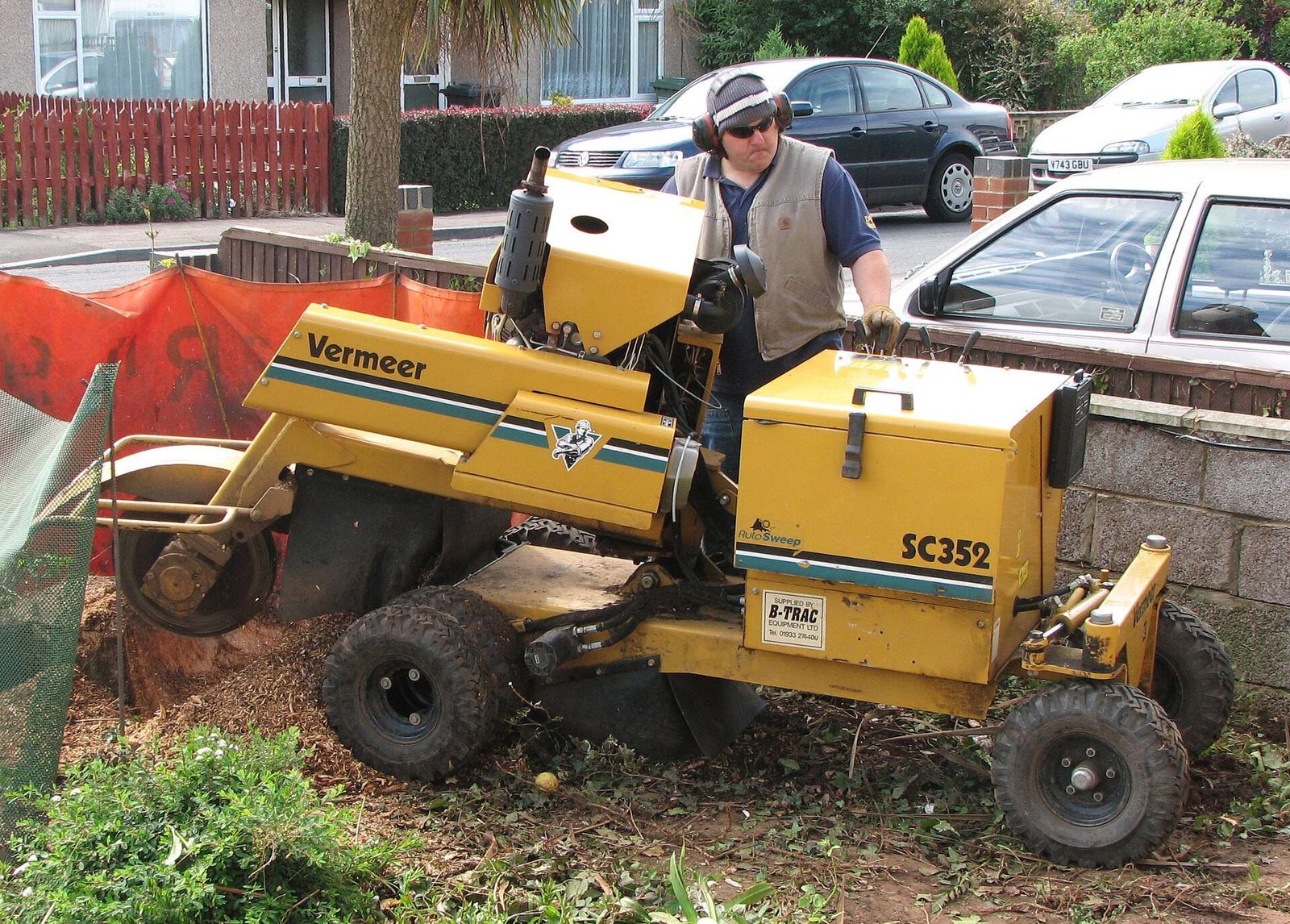 Operator guiding a walk-behind stump grinder over a stump beside a residential street